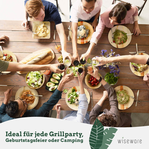 Eine Gruppe stößt mit ihren Gläsern gemeinsam über einem Tisch an, verschiedene Pastagerichte und Beilagen sind auf Palmblatttellern und Palmblattschalen serviert.